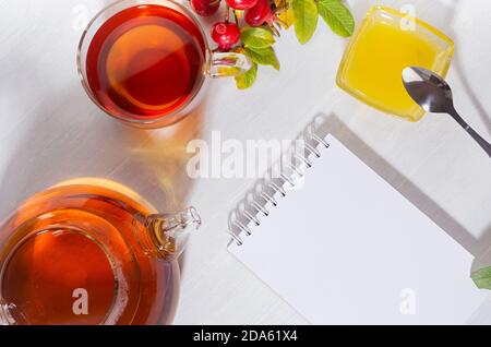 Thé d'automne confortable avec thé aux hanches roses dans une théière avec tasse, branche de rosier de chien et carnet vierge avec ombres sur la lumière douce, vue sur le dessus de table en bois blanc, cop Banque D'Images