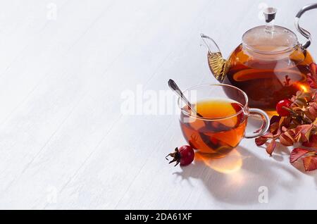 Agréable thé d'automne matinal avec thé aux hanches roses dans une théière avec tasse, branche de rose de chien avec des rayons de soleil et des ombres sur la lumière douce de la table en bois blanc vue sur le dessus, c Banque D'Images