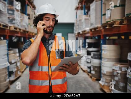 Le gestionnaire d'entrepôt suit les détails de la commande d'approvisionnement à l'aide d'une tablette numérique lors d'une conversation sur un téléphone mobile dans l'entrepôt Banque D'Images