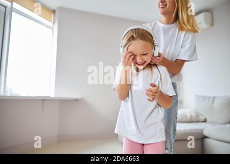 Une petite femme souriante écoute de la musique dans les dépenses d'écouteurs temps avec maman dans la chambre à l'intérieur Banque D'Images
