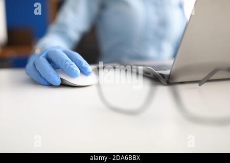 Main dans le gant médical bleu repose sur la souris d'ordinateur à côté aux lunettes et à l'ordinateur portable Banque D'Images