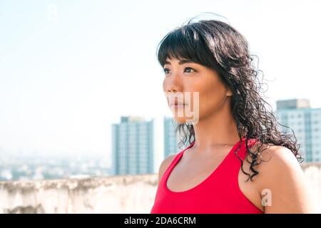 Jeune femme asiatique regardant loin dans les gratte-ciel sur les toits. Athelete féminin déterminé dans la tenue de sport avec espace de copie Banque D'Images