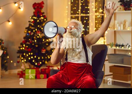 Le Père Noël senior parle dans un haut-parleur tout en étant assis dans une pièce avec des décorations de Noël. Banque D'Images