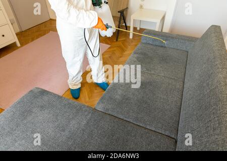 Personne en combinaison de protection avec un pulvérisateur de décontamination désinfectant le ménage et le mobilier. Banque D'Images