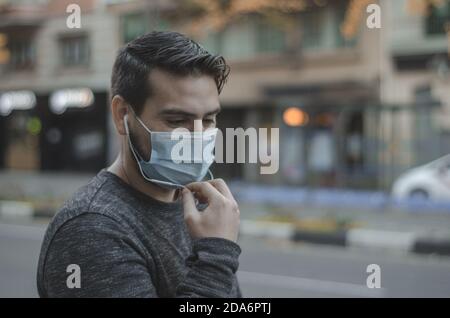 Jeune homme portant un masque, parlant au téléphone et attendant son bus pour arriver Banque D'Images