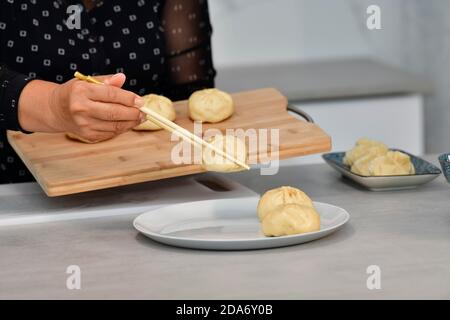 Gros plan d'une main femelle utilisant des baguettes pour déplacer des petits pains chinois traditionnels d'une planche à découper à une plaque. Concept de la cuisine chinoise traditionnelle. Banque D'Images
