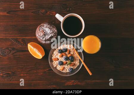 Petit déjeuner équilibré avec bol de flocons d'avoine, baies, noix, dessert et une tasse de café sur une table de cuisine brune, vue sur le dessus. Banque D'Images
