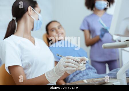 Jeune femme médecin en uniforme, gants en caoutchouc et masque avec outil traite dent de petit enfant souriant Banque D'Images