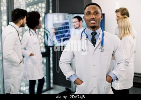 Jeune beau médecin afro-américain souriant avec ses collègues multiraciaux, debout à l'arrière-plan, ayant chat vidéo avec un médecin masculin Banque D'Images