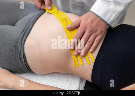 Kinésithérapeute mâle appliquant du ruban élastique thérapeutique adhésif jaune sur la femelle muscle abdominal de jeune adulte mince femme sportive couché table de traitement Banque D'Images