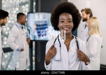 Souriant jolie infirmière afro-américaine ou médecin, posant à la caméra en clinique. Équipe médicale multiraciale discutant en arrière-plan, discutant en ligne Banque D'Images