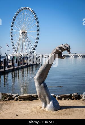 Une sculpture d'un bras dépasse du sable devant la roue de la capitale à National Harbor, Maryland, avec le pont Woodrow Wilson derrière. Banque D'Images