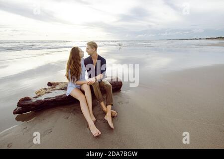 Couple assis sur un tronc d'arbre mort sur une plage à Bali au coucher du soleil, regardant l'un l'autre Banque D'Images