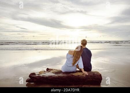 Couple assis sur un tronc d'arbre mort sur une plage À Bali au coucher du soleil Banque D'Images