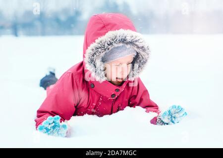 Drôle caucasien souriant fille dans chaud hiver vêtements rose veste jouant avec la neige. Enfant mignon couché sur le sol pendant la journée froide d'hiver enneigée à la chute de neige Banque D'Images