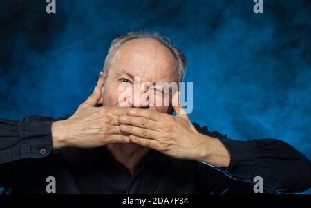 Vieux macho. Un homme âgé couvre sa bouche avec ses paumes. Portrait d'un homme émotionnel âgé sur fond sombre dans le studio Banque D'Images