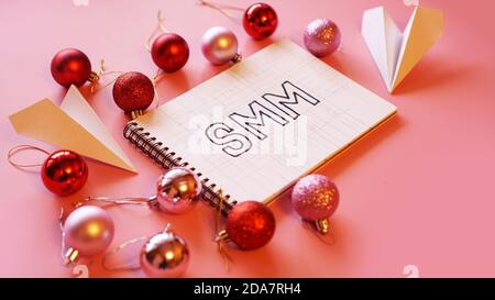 L'inscription SMM dans un carnet sur un fond rose festif. Boules de Noël rouges et roses, avions en papier autour Banque D'Images