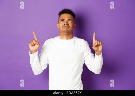 Déçu et triste afro-américain jeune gars en sweat-shirt blanc, regardant et pointant les doigts vers le haut, montrant quelque chose de mauvais, en se portant bouleversé par le violet Banque D'Images