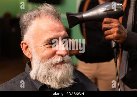 Les jeunes Afro-américains tatoués peignes de barbier et de souffle-drys les cheveux de Homme caucasien mature avec une barbe blanche Banque D'Images