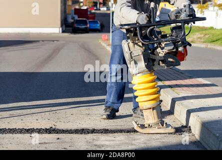 Un ouvrier de la route utilise un rameur portatif de vibrations d'essence pour réparer l'asphalte sur l'allée lors d'une journée ensoleillée. Banque D'Images