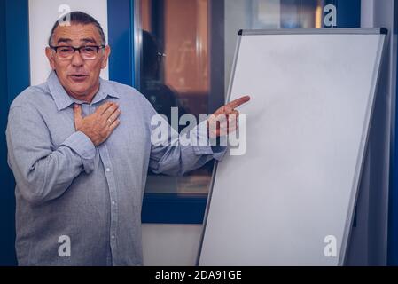 un homme adulte avec des lunettes et fait une présentation de idées devant un tableau noir touchant sa poitrine Banque D'Images