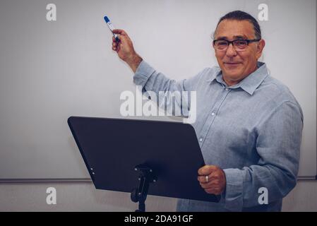 un homme adulte avec des lunettes fait une présentation sur un lutrin et un tableau noir en arrière-plan avec un stylo en main Banque D'Images