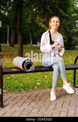 Portrait de Smiling Fitness jeune femme assise sur un banc dans le parc de la ville et portant des écouteurs. Banque D'Images