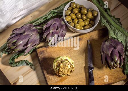 Recette sicilienne typique : un coeur d'artichaut farci, quelques artichauts violets Tema et un bol avec des olives Nocellara Banque D'Images