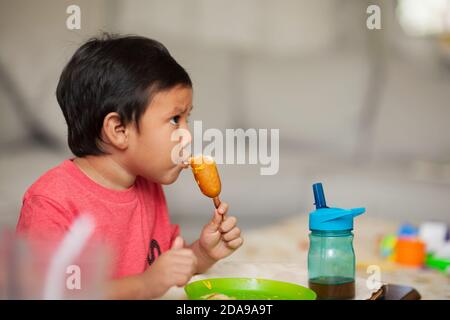 Un petit enfant qui regarde un spectacle tout en prenant un repas sain pour le déjeuner, en mangeant un chien de maïs avec du jus de raisin. Banque D'Images