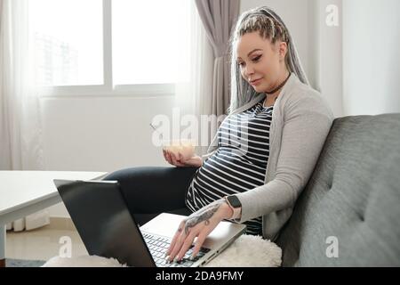 Belle jeune femme enceinte qui mange de la salade de fruits en regardant un film ou blog sur ordinateur portable Banque D'Images