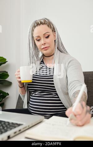 Une jeune femme enceinte boit une tasse de café sans caféine, travaille sur un ordinateur portable et prend des notes dans le planificateur Banque D'Images