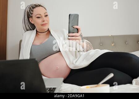 Belle jeune femme enceinte heureuse se reposant sur le lit et lisant publications sur les médias sociaux Banque D'Images