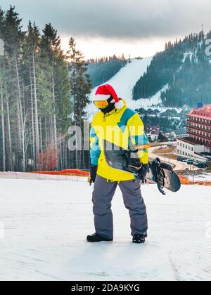 Homme avec snowboard à red christmas hat walking up par neigé hill Banque D'Images