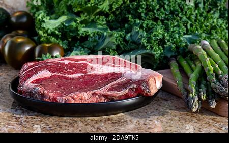 Viande crue, steak de bœuf sur une assiette noire avec chou vert, asperges, broccolini et tomates en arrière-plan. Banque D'Images