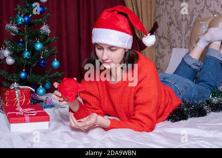 Une belle jeune fille économise de l'argent pour Noël et le nouvel an. Une jeune fille dans un chapeau de Noël et un chandail rouge tire les pièces d'une banque de piggy rouge à Banque D'Images