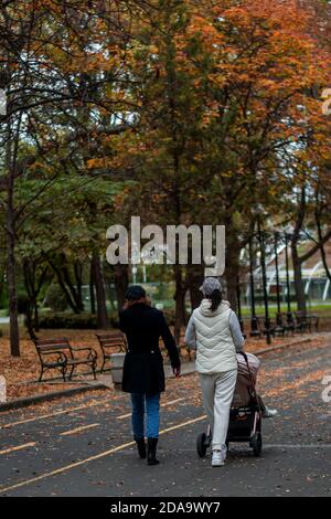 Une mère marche avec une poussette et une deuxième femme sur une route dans un parc pendant la saison d'automne. Deux femmes marchant. Banque D'Images