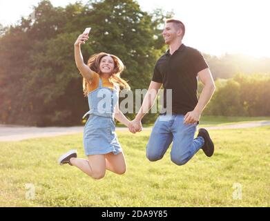 Heureux couple millénaire sautant ensemble et emportant le selfie à l'extérieur Coucher de soleil - concept de loisirs dans un parc avec jeunes heureux amis Banque D'Images