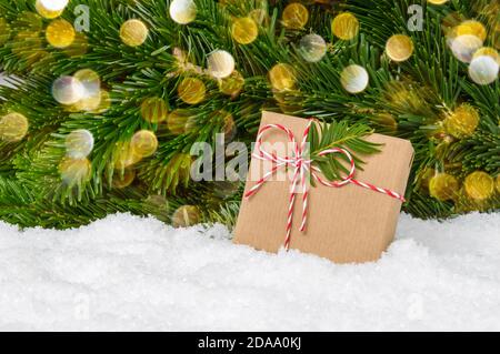 Boîte cadeau de Noël faite à la main sous un sapin. Présent enveloppé dans du papier artisanal et des supports d'arc de corde dans la neige avec des branches de pin et des lumières de bokeh comme bac Banque D'Images