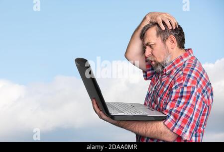 Problèmes avec l'ordinateur. Homme d'affaires d'âge moyen stressé travaillant sur un ordinateur portable contre le ciel bleu avec des nuages Banque D'Images