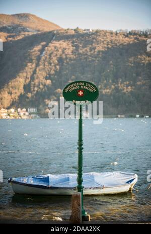 Panneau sur le front de mer et un bateau sur le lac alpin de Lugano avec montagne en une journée ensoleillée à Morcote, Tessin en Suisse. Banque D'Images