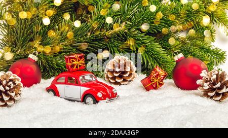 Fulda, Allemagne - 24 octobre 2020 : voiture rouge dans la neige avec des cônes de pin, des boules de Noël, des cadeaux, des sapins et des lumières de bokeh. Vacances d'hiver ou delive Banque D'Images