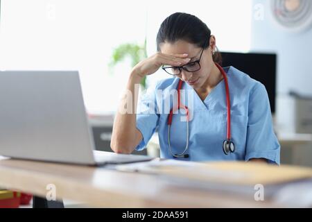 Jeune femme médecin assis à la table et tenant la tête dedans avant de l'ordinateur portable dans le bureau Banque D'Images