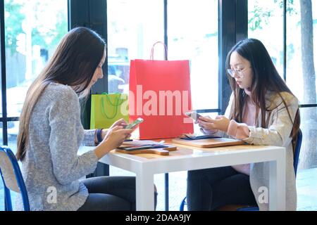 Deux amies dans un café-restaurant qui vérifient leur smartphone après avoir fait du shopping. Les femmes caucasiennes et asiatiques dans un restaurant. Banque D'Images