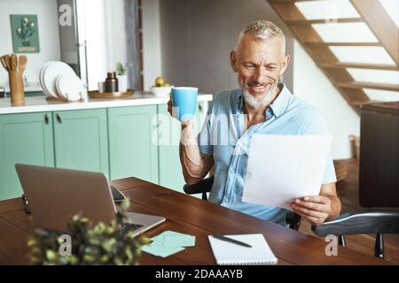 Joyeux homme de race blanche d'âge moyen assis à table avec du café tasse pendant la journée de travail à distance à la maison Banque D'Images