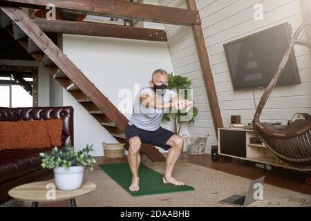 Rester actif dans l'isolement. Homme sportif d'âge moyen portant un masque de protection du visage s'exerçant à la maison tout en regardant l'entraînement en ligne sur un ordinateur portable Banque D'Images
