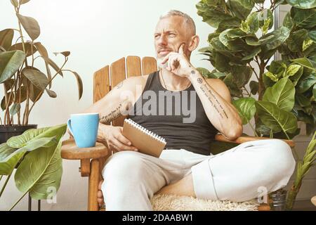 Perdu dans les pensées. Homme d'âge moyen concentré assis sur la terrasse ensoleillée de son appartement moderne, tenant son carnet et pensant à quelque chose Banque D'Images
