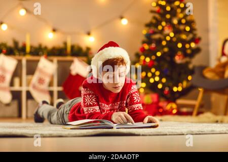 Un garçon concentré dans des vêtements de Noël rouges et un chapeau allongé étage et livre de lecture Banque D'Images