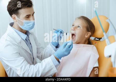 Jeune homme de soin attrayant en manteau blanc, masque de protection et gants faisant procédure médicale pour le traitement dentaire Banque D'Images