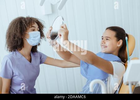 Bonne petite fille regarde dans le miroir pour estimer le travail de dentiste et profitez du résultat Banque D'Images