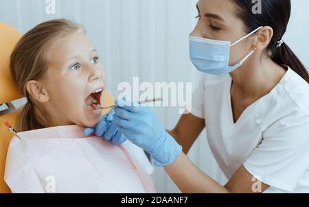 Jeune femme sérieuse attrayante en manteau blanc, masque de protection et gants en caoutchouc examine les dents de la petite fille Banque D'Images